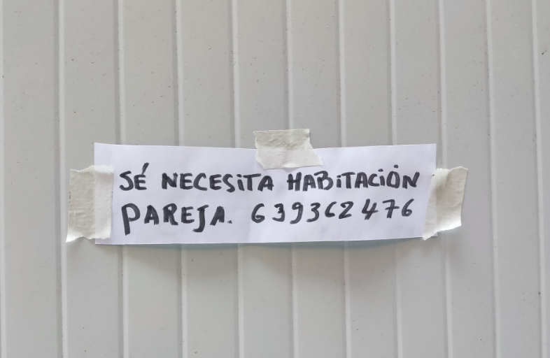 Cartel callejero con el texto "SÉ NECESITA HABITACIÓN PAREJA." y un número